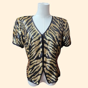 May include: A short-sleeved blouse covered in black and gold sequins arranged in a striped pattern. The garment features a V-neckline, a front opening, and a structured shoulder design, displayed on a mannequin to show the overall fit and shape.