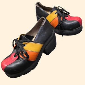May include: A pair of black platform shoes with a colorful design. The toe is red, the middle section is yellow and orange, and the laces are black. The shoes have a chunky heel and the word "DESTROY" is visible on the side.