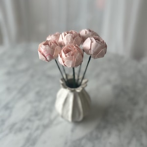 Può includere: Un bouquet di sei fiori di carta rosa pallido con petali piegati e stratificati attaccati a sottili steli scuri. I fiori sono disposti in un piccolo vaso in ceramica bianca scanalata con bordo smerlato, creando una composizione floreale strutturata.