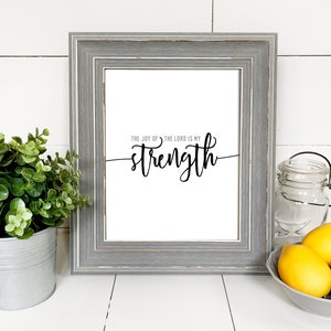 Puede incluir: Una impresión enmarcada con el texto "The joy of the Lord is my strength" en escritura cursiva negra sobre fondo blanco. El marco es gris con un acabado desgastado. La impresión está sobre una superficie blanca con una planta en maceta y limones en un cuenco.