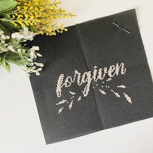 Peut inclure: Une toile noire avec le mot "forgiven" brodé en fil blanc. La toile est sur une surface blanche avec un bouquet de fleurs jaunes et blanches.
