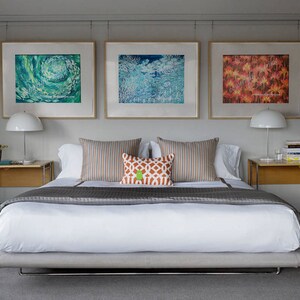 May include: A white bed with a gray bedspread and a white headboard. The bed is in a bedroom with three framed pictures on the wall above the headboard. The pictures are of abstract art in shades of blue, green, and orange.
