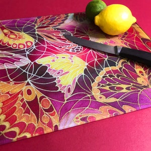 May include: A glass chopping board with a colourful butterfly design. The board features a purple, red and gold pattern with intricate details. A knife is placed on the board.