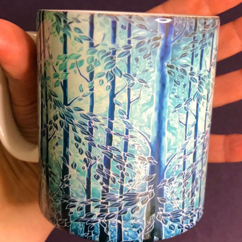 May include: A white ceramic mug with a blue and green forest scene design. The design features stylized trees and leaves in a watercolor style.