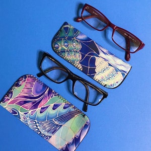 Op de afbeelding: Twee brillen met gepatroneerde stoffen etuis. De brillen zijn beide zwart en hebben een rechthoekige vorm. Een etui is blauw met een wit en groen bloemenpatroon. Het andere etui is blauw met een wit en geel schelpmotief.