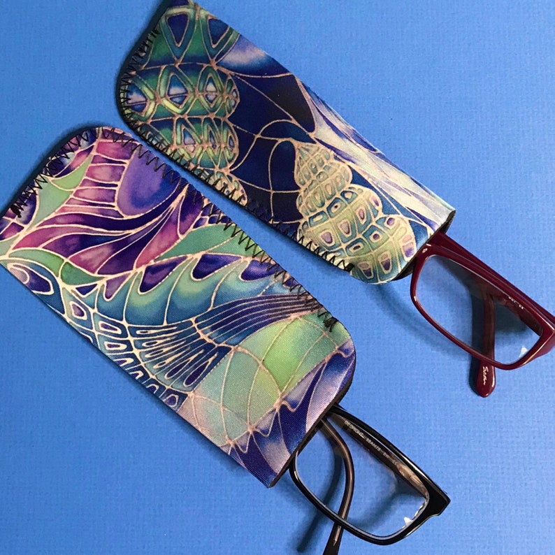 Op de afbeelding: Twee stoffen brillenkokers met een kleurrijk abstract ontwerp. De ene koker is blauw en paars, de andere is groen en blauw. Beide kokers hebben een zigzagsteek rond de randen.