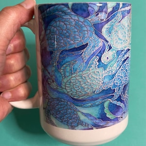 Könnte beinhalten: Weiße Keramiktasse mit einem blauen und silbernen Design von Meeresschildkröten. Die Schildkröten überlappen sich und haben einen Glasmalerei-Look.