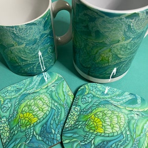 Könnte beinhalten: Zwei weiße Keramikbecher mit einem abstrakten Design in Grün und Blau. Die Becher stehen auf zwei passenden quadratischen Untersetzern mit dem gleichen Design.
