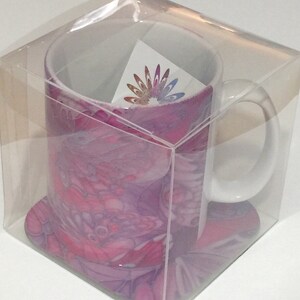 K&ouml;nnte beinhalten: Eine wei&szlig;e Keramiktasse mit einem abstrakten Design in Rosa und Lila. Die Tasse ist in einer durchsichtigen Plastikbox mit einem passenden Untersetzer verpackt.