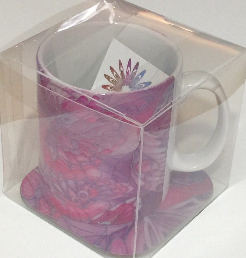 K&ouml;nnte beinhalten: Eine wei&szlig;e Keramiktasse mit einem abstrakten Design in Rosa und Lila. Die Tasse ist in einer durchsichtigen Plastikbox mit einem passenden Untersetzer verpackt.
