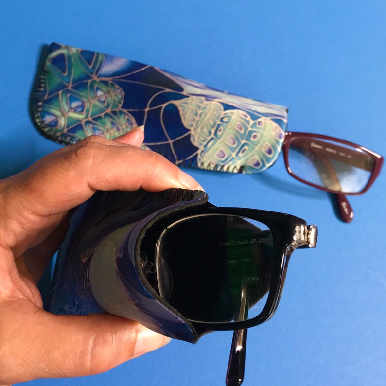 Op de afbeelding: Een zwarte zonnebril met een donker glas en een zilveren montuur. De zonnebril zit in een blauwe en groene bedrukte etui. De etui heeft een ritssluiting.