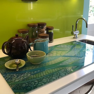May include: A brown teapot, a blue jug, and a green bowl with a spoon are on a green and blue patterned glass chopping board. The chopping board is on a white worktop. There are five glass jars with wooden lids behind the teapot and jug.