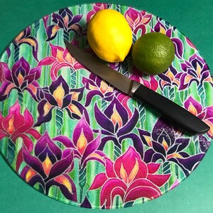 May include: A round glass chopping board with a floral design featuring purple, pink, and yellow irises on a green background. A black handled knife, a yellow lemon, and a green lime are on the board.