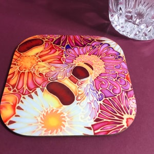 May include: Square coaster with a floral design in shades of orange, pink, and purple. The coaster features a detailed pattern of stylized flowers and leaves, with a dark brown border. A clear glass is partially visible in the background.