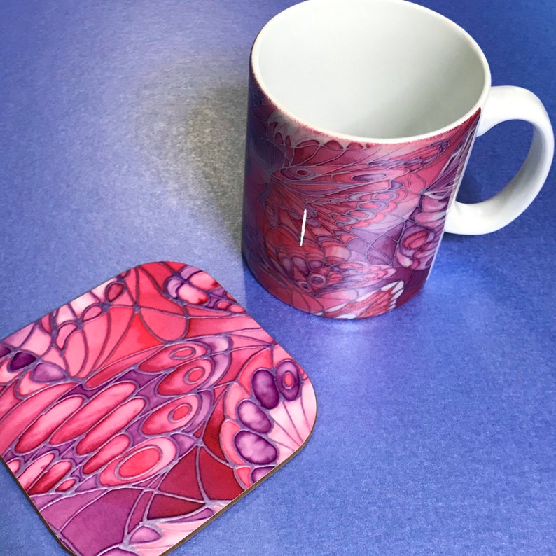 K&ouml;nnte beinhalten: Eine rosa und lila Keramiktasse mit einem Schmetterlingsdesign. Die Tasse steht auf einem Untersetzer mit passendem Design.