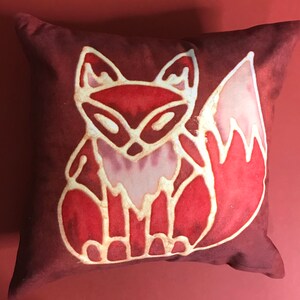 May include: A decorative cushion featuring a stylised fox design in red and white. The fox is depicted sitting with its tail curled around its body. The cushion is made from a soft, plush fabric.