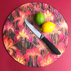 May include: A round glass chopping board with a floral design in shades of pink, red, yellow, and green. The board features a knife and two pieces of fruit, a lime and a lemon.
