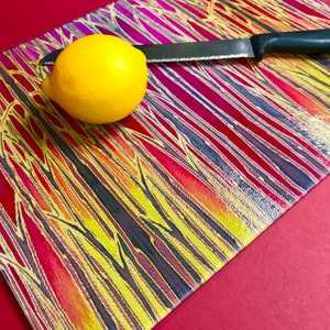 May include: A yellow lemon sits on a colourful chopping board with a silver knife. The chopping board has a striped pattern with red, yellow, pink, and purple hues.