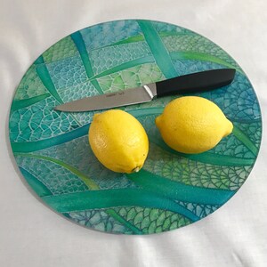 May include: A round glass chopping board with a green and blue mosaic design. Two yellow lemons and a silver knife are on the board.