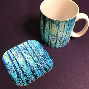 May include: A white ceramic mug with a blue and green forest design. The mug is paired with a matching coaster.