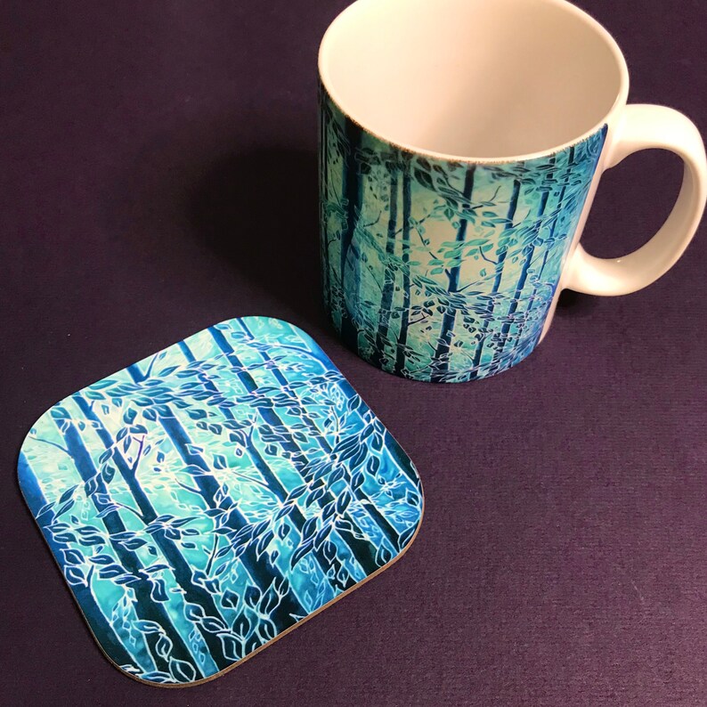 May include: A white ceramic mug with a blue and green forest design. The mug is paired with a matching coaster.