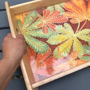 May include: A wooden serving tray featuring a colourful leaf design. The tray has a light wood frame and a clear, glossy finish. The leaves are painted in shades of red, orange, yellow, and green.