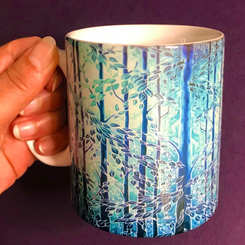 May include: A white ceramic mug with a blue and green forest scene printed on it. The scene features trees with leaves and a light blue background.