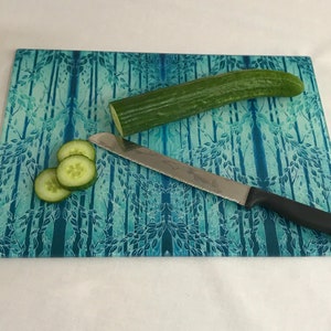 May include: A blue glass chopping board with a forest design features a cucumber and a knife. The cucumber is sliced and the knife is lying on the board.
