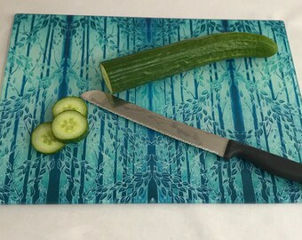 Op de afbeelding: Een blauw glazen snijplank met een bosontwerp heeft een komkommer en een mes. De komkommer is in plakjes gesneden en het mes ligt op het plank.