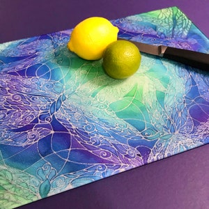 May include: A glass chopping board with a blue, green and purple watercolour design featuring white swirls. A yellow lemon and a green lime are on the board.