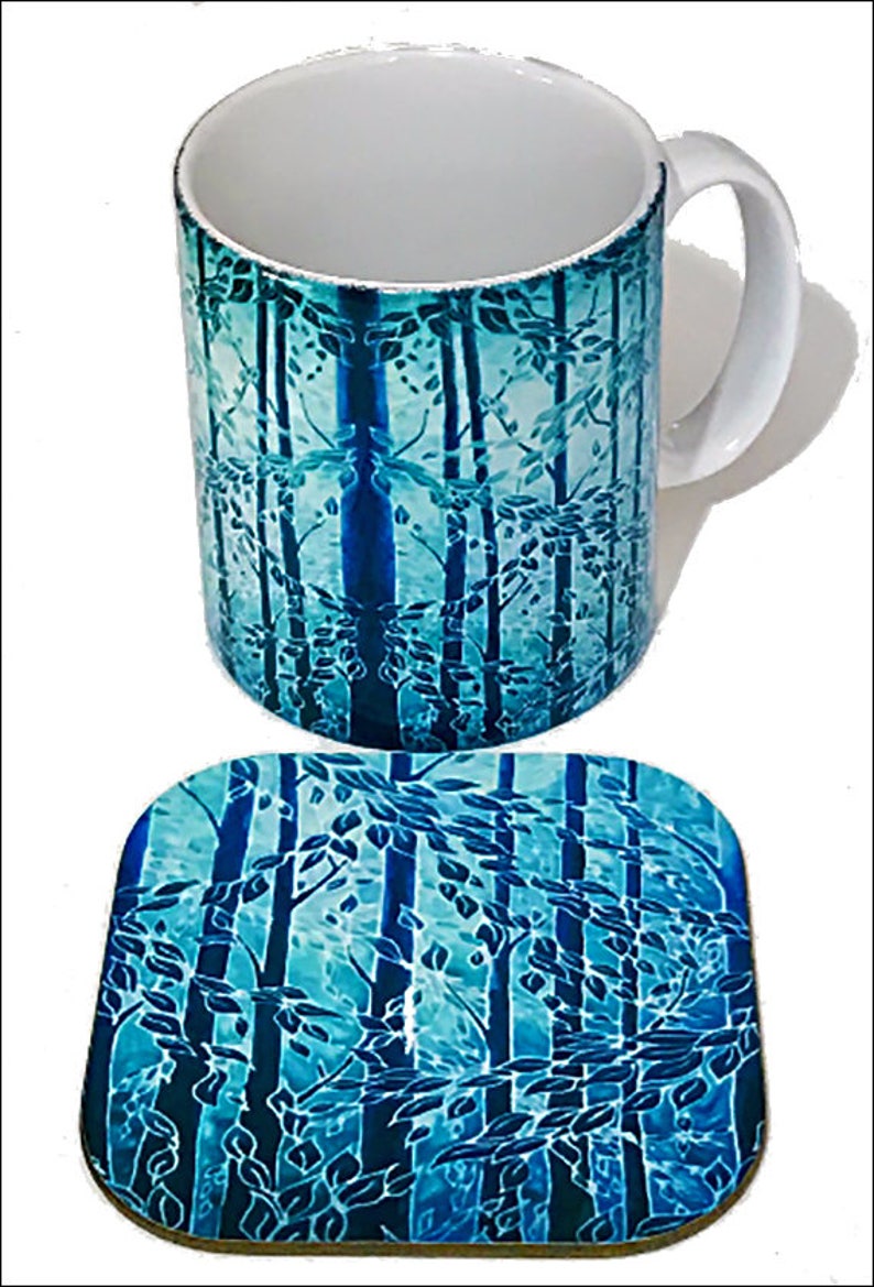 May include: A blue and green ceramic mug and matching coaster with a forest scene design. The mug has a white interior and a handle. The coaster is square shaped.
