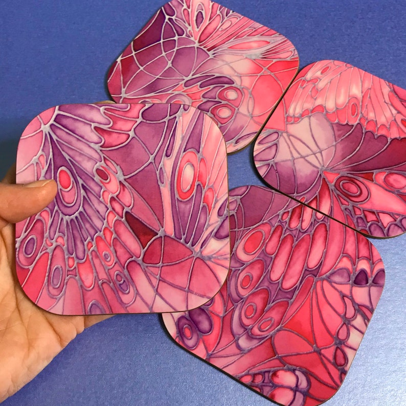 K&ouml;nnte beinhalten: Satz mit vier quadratischen Untersetzern mit einem Design aus rosa und violetten Schmetterlingsfl&uuml;geln. Die Untersetzer sind aus Holz und haben eine gl&auml;nzende Oberfl&auml;che.