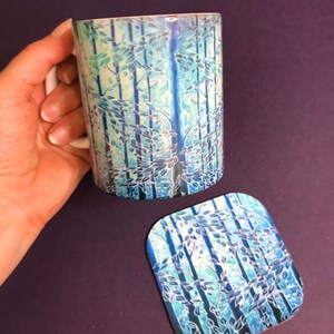 May include: A white ceramic mug with a blue and green forest design. The mug has a handle and is sitting on a matching coaster.