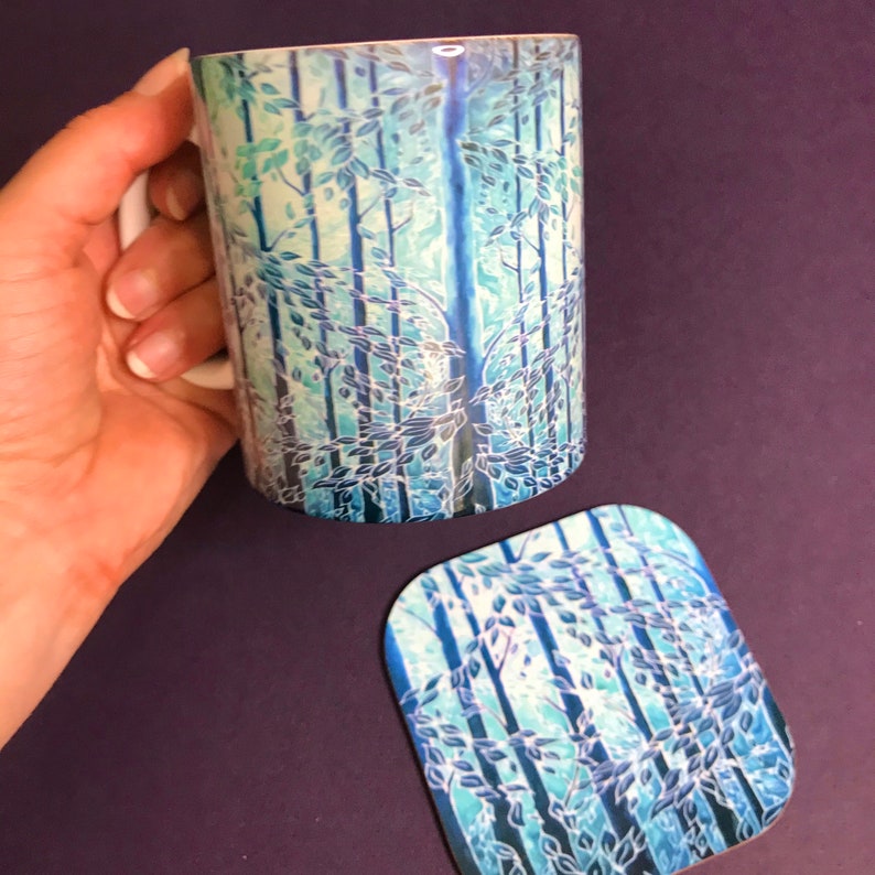 May include: A white ceramic mug with a blue and green forest design. The mug has a handle and is sitting on a matching coaster.