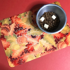 Puede incluir: Un cuenco de metal plateado para perros lleno de comida para perros marr&oacute;n y golosinas para perros blancas se encuentra sobre un mantel individual estampado en rojo y naranja con un dise&ntilde;o de hojas.