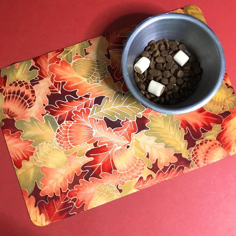 Puede incluir: Un cuenco de metal plateado para perros lleno de comida para perros marr&oacute;n y golosinas para perros blancas se encuentra sobre un mantel individual estampado en rojo y naranja con un dise&ntilde;o de hojas.