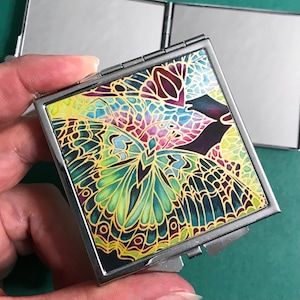 Può includere: Uno specchio compatto argentato con un design a farfalla colorato. Le ali della farfalla sono un mix di verde, giallo, blu e viola.
