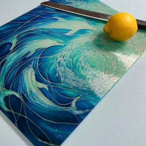 May include: A blue and green glass chopping board with a swirling pattern of dolphins and waves. A lemon and a knife are on the board.