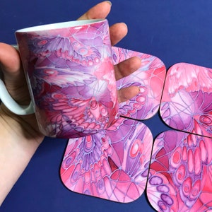 K&ouml;nnte beinhalten: Ein Set aus vier Untersetzern in Rosa und Lila mit einem Schmetterlingsfl&uuml;gel-Design. Die Untersetzer sind aus Keramik und haben eine gl&auml;nzende Oberfl&auml;che. Eine Tasse mit dem gleichen Design ist ebenfalls abgebildet.