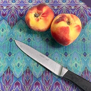 May include: Two ripe peaches, red and yellow, on a decorative cutting board with a teal and purple floral design. A stainless steel knife with a black handle is also on the board.
