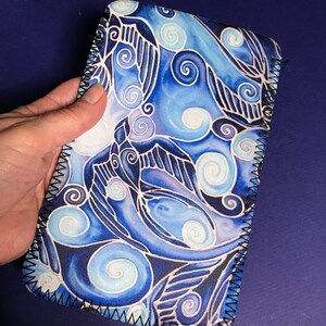 May include: Blue and white fabric phone case with a swirling pattern of birds and clouds. The case has a black stitched edge.