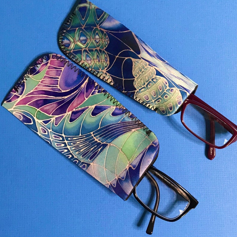 Op de afbeelding: Twee gepatroneerde brillenetuis van stof met brillen erin. De etuis zijn blauw, groen en paars met een draaiend ontwerp. Een etui bevat een rood brilletje en het andere een zwart brilletje.