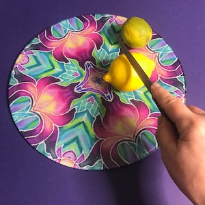May include: A round glass chopping board with a colourful floral design. A yellow lemon and a green lime are on the board, and a silver knife is being used to cut the lemon.
