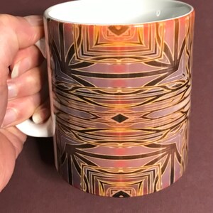 May include: A white ceramic mug with a geometric pattern in shades of brown, gold, and purple. The pattern is symmetrical and features intricate lines and shapes.