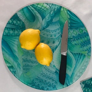 May include: A round glass serving tray with a teal and green abstract design. Two lemons and a silver knife are on the tray.