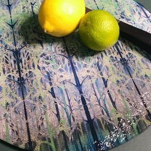 May include: A round glass chopping board with a blue and green forest design. A yellow lemon and a green lime are on the board.