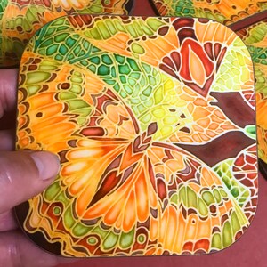 May include: A square coaster with a colorful butterfly design. The butterfly's wings are made up of intricate patterns in shades of orange, yellow, green, and brown.