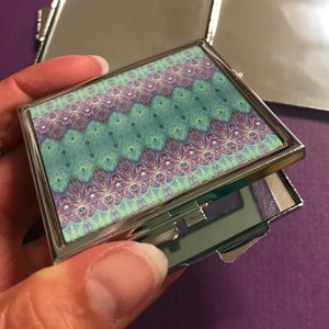 Può includere: Uno specchio compatto argentato con un coperchio incernierato. Il coperchio presenta un motivo geometrico colorato in tonalità di viola, blu e verde.