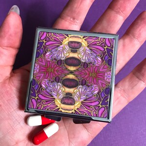 May include: A small, square pill box with a hinged lid. The box is decorated with a colorful, intricate design of purple, gold, and pink flowers and butterflies. Two white and red capsules are lying on a hand in front of the box.