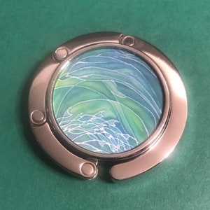 May include: A silver-toned metal purse hook with a round, green and blue patterned design. The design features abstract swirls and lines.